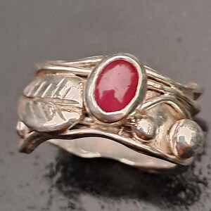 Könnte beinhalten: Ein silberner Ring mit einem roten ovalen Edelstein, der in einem verdrehten Band gefasst ist. Das Band ist mit Blatt- und Beerenmotiven verziert.