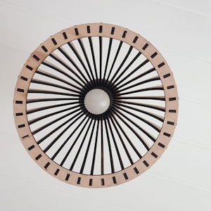 May include: A circular pendant light fixture featuring a light-colored wooden outer ring and a series of dark, thin, radiating spokes that converge toward a central white light bulb, creating a geometric, sunburst-like pattern.