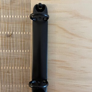 May include: Black metal binder clip with two rings. The clip is on a light wood surface.