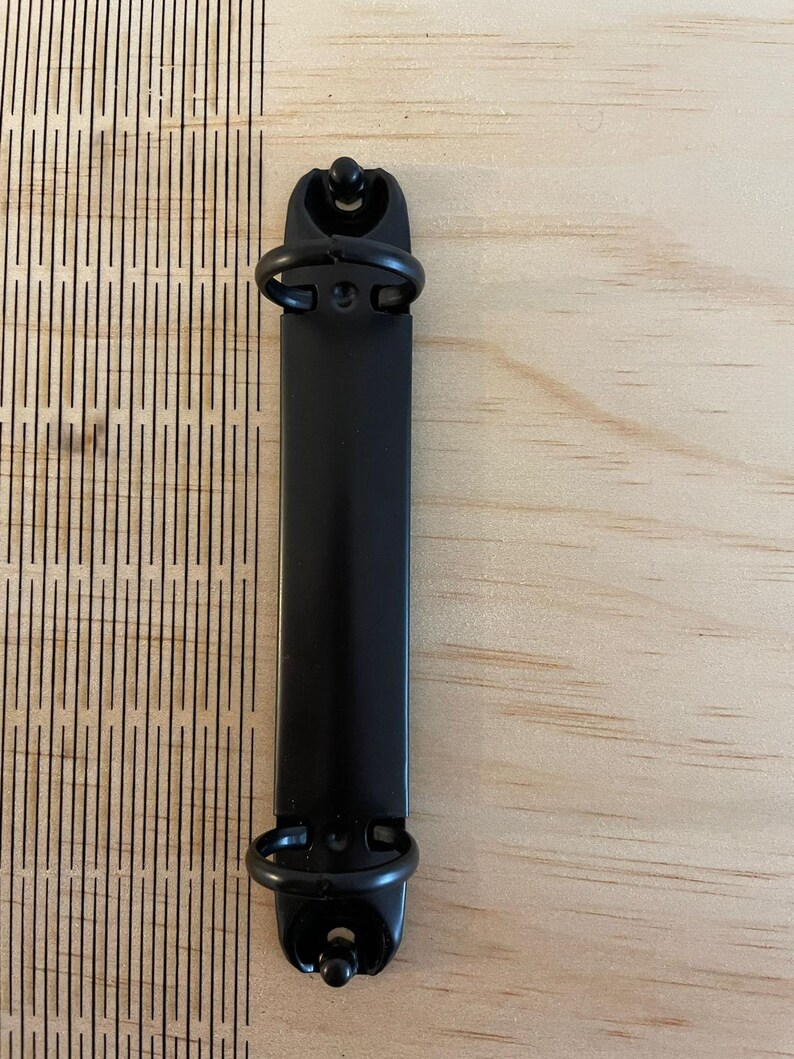 May include: Black metal binder clip with two rings. The clip is on a light wood surface.
