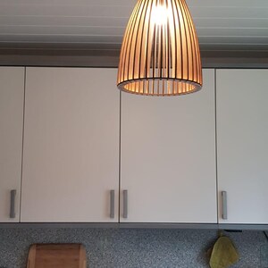 May include: A pendant light fixture featuring a cylindrical shade constructed from vertical dark brown wooden slats. The light is suspended from a black cord and ceiling mount, emitting a warm glow from the internal bulb, illuminating the kitchen.