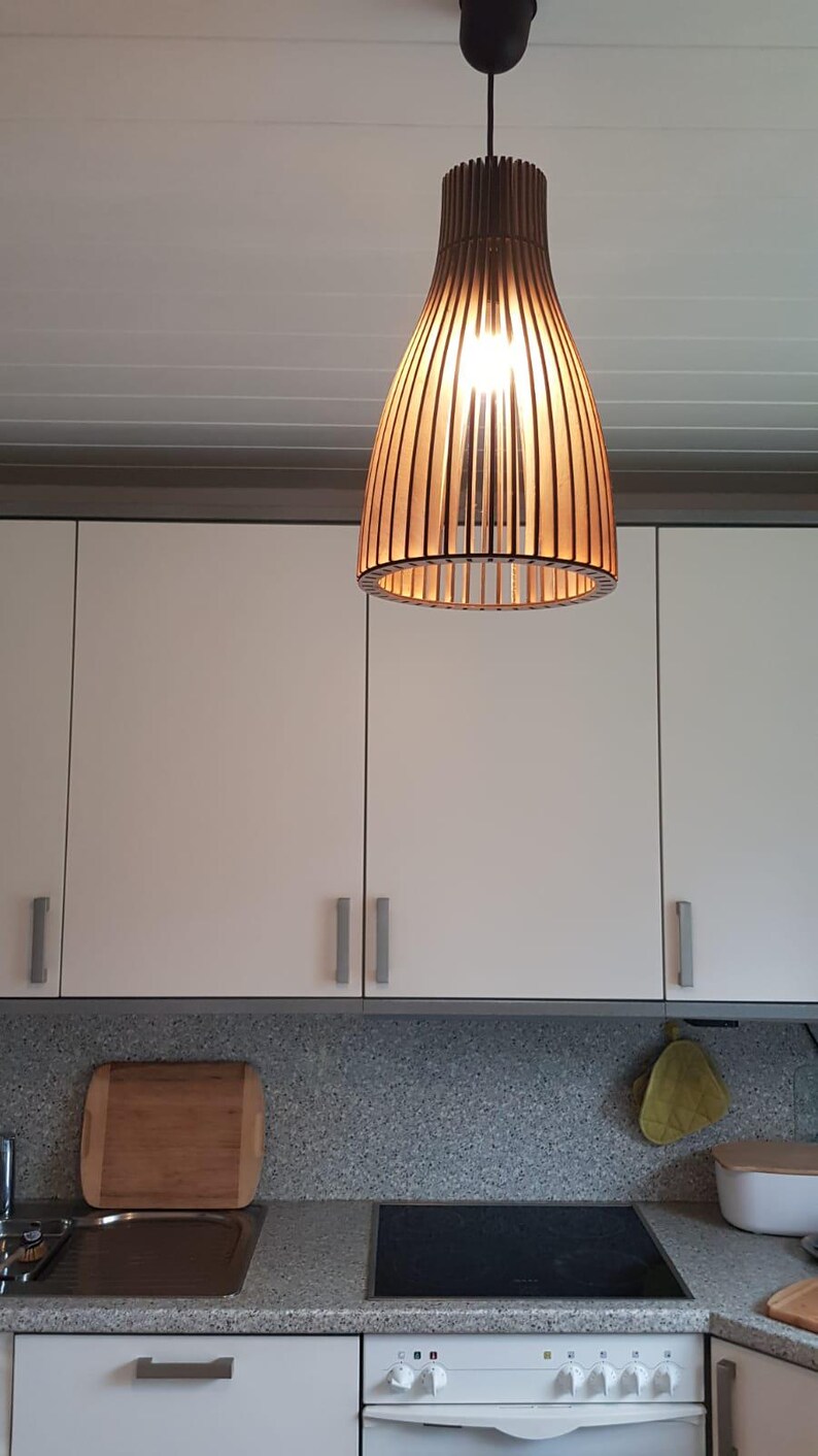 May include: A pendant light fixture featuring a cylindrical shade constructed from vertical dark brown wooden slats. The light is suspended from a black cord and ceiling mount, emitting a warm glow from the internal bulb, illuminating the kitchen.