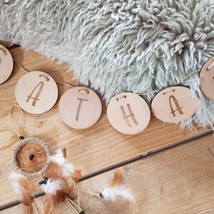 May include: A wooden banner spelling out the name "NATHA" in capital letters. Each letter is on a separate, round wooden disc. The banner is strung on a thin, natural-coloured cord. A small dreamcatcher with feathers hangs below the banner, on a wooden surface.