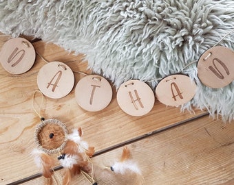 May include: A wooden banner spelling out the name "NATHA" in capital letters. Each letter is on a separate, round wooden disc. The banner is strung on a thin, natural-colored cord. A small dreamcatcher with feathers hangs below the banner, on a wooden surface.