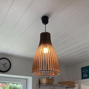 May include: A pendant light fixture featuring a slatted wooden shade with a bell-shaped silhouette. The lamp is suspended from a black ceiling mount and emits a warm glow from a central light bulb, casting light through the vertical wooden slats.