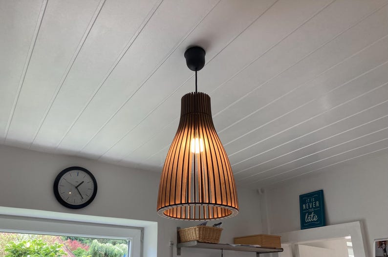 May include: A pendant light fixture featuring a slatted wooden shade with a bell-shaped silhouette. The lamp is suspended from a black ceiling mount and emits a warm glow from a central light bulb, casting light through the vertical wooden slats.