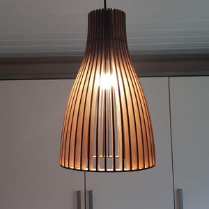 May include: A bell-shaped pendant light fixture constructed from vertical dark wood slats. The lamp features a black ceiling mount and cord, with a warm light source visible through the open gaps between the wooden slats, creating a linear pattern.