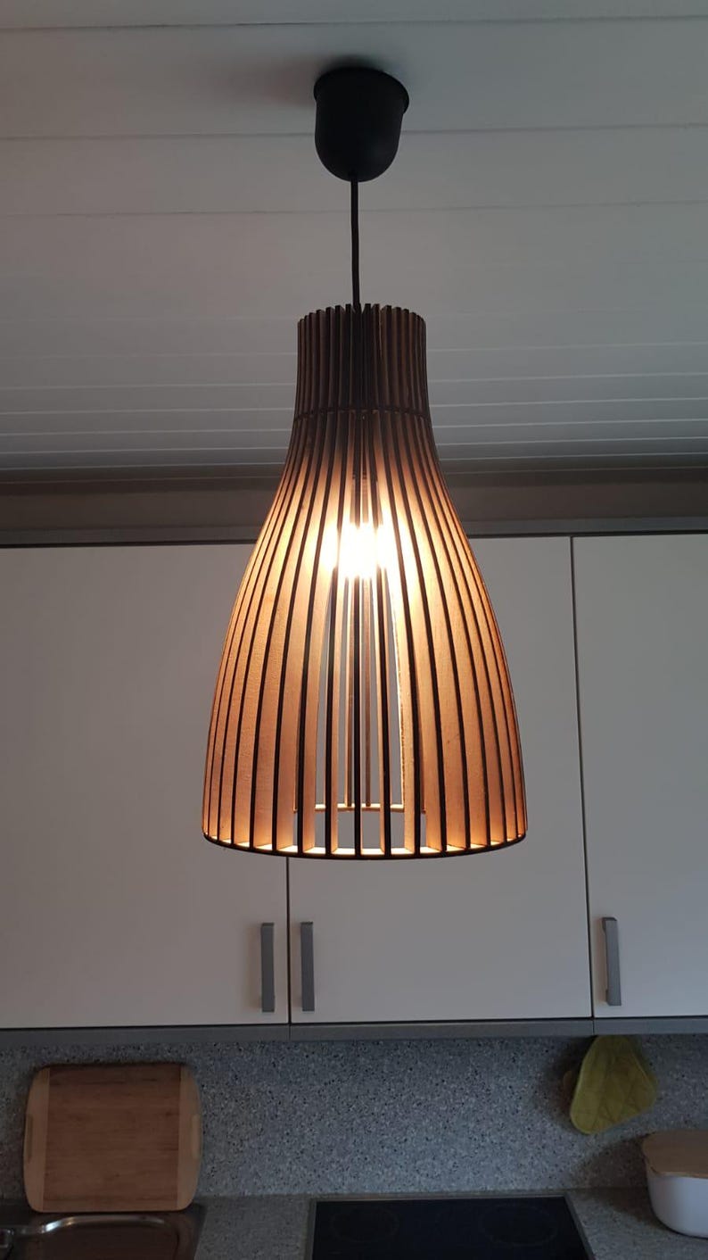 May include: A bell-shaped pendant light fixture constructed from vertical dark wood slats. The lamp features a black ceiling mount and cord, with a warm light source visible through the open gaps between the wooden slats, creating a linear pattern.