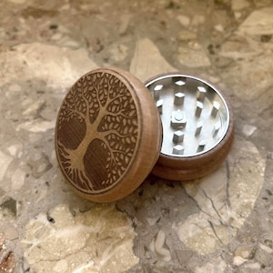 May include: A wooden herb grinder with a tree of life design. The top piece is engraved with the tree image, while the bottom piece has a metal grinding surface. The grinder is made of light brown wood.