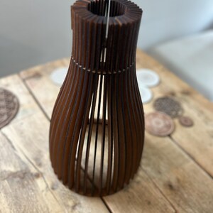 May include: A dark brown wooden vase constructed from a series of vertical slats arranged in a rounded, open-frame silhouette. The structure features a narrow neck and a wider, curved body, creating a hollow, cage-like appearance with visible gaps.