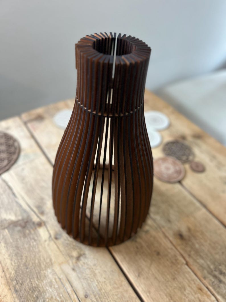 May include: A dark brown wooden vase constructed from a series of vertical slats arranged in a rounded, open-frame silhouette. The structure features a narrow neck and a wider, curved body, creating a hollow, cage-like appearance with visible gaps.