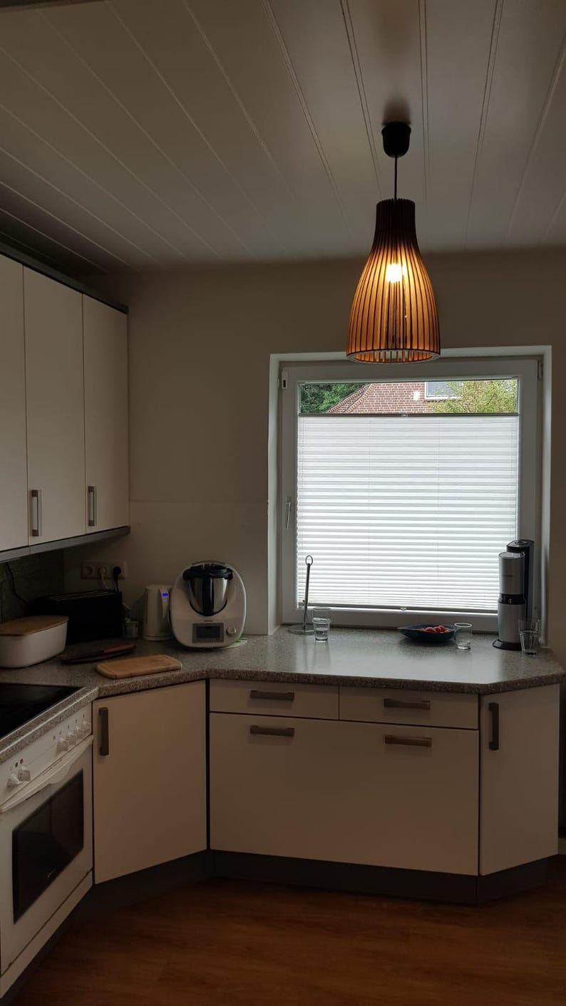 May include: A kitchen interior featuring white cabinets with silver handles, a speckled gray countertop, and a wooden slatted pendant light hanging above. A window with white pleated blinds, a food processor, and a toaster are visible.