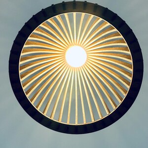 May include: A circular pendant light fixture featuring a dark outer ring frame with numerous thin, light-colored wooden slats radiating inward toward a central, glowing white light bulb, creating a sunburst pattern against a pale background.