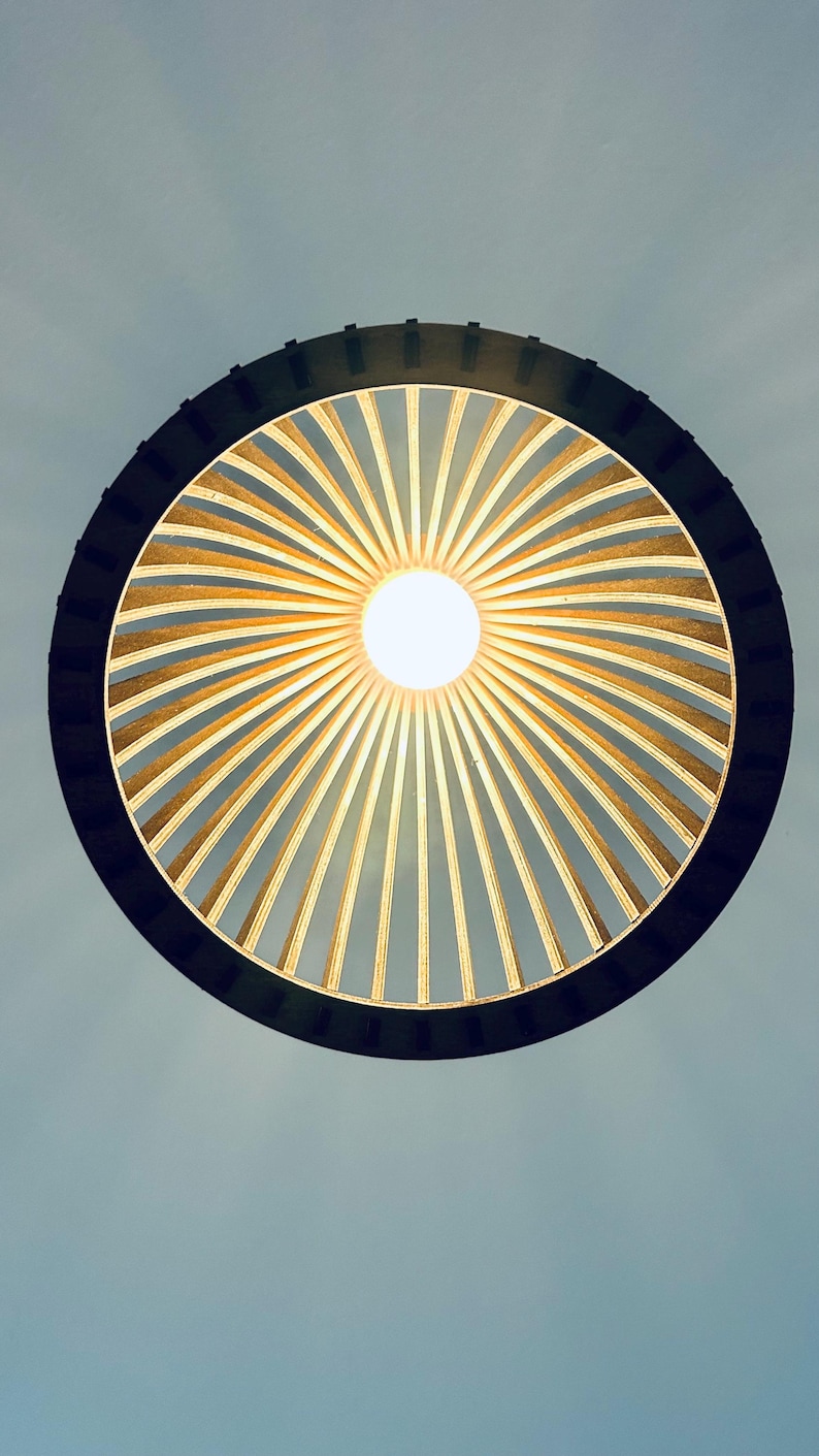 May include: A circular pendant light fixture featuring a dark outer ring frame with numerous thin, light-colored wooden slats radiating inward toward a central, glowing white light bulb, creating a sunburst pattern against a pale background.