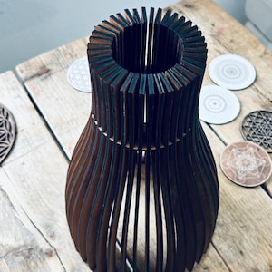 May include: A dark brown wooden vase constructed from vertical slats arranged in a circular, open-frame pattern. The structure features a narrow neck that flares outward toward the base, creating a hollow, cage-like silhouette with visible gaps.