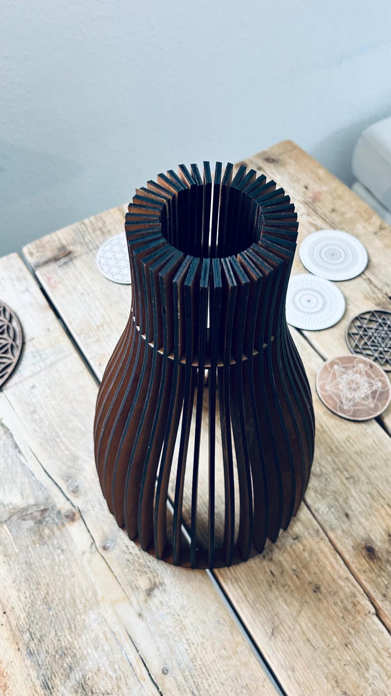 May include: A dark brown wooden vase constructed from vertical slats arranged in a circular, open-frame pattern. The structure features a narrow neck that flares outward toward the base, creating a hollow, cage-like silhouette with visible gaps.