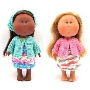 May include: Two crocheted dolls wearing matching dresses and cardigans. One doll has brown hair and a light blue cardigan, the other has blonde hair and a pink cardigan.