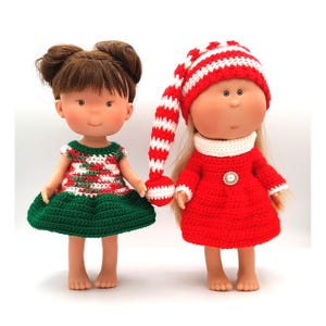 Może przedstawiać: Dwie lalki ubrane w ręcznie robione szydełkowe stroje. Jedna lalka ma na sobie zielono-czerwoną i białą sukienkę. Druga lalka jest w czerwonej sukience i długiej, pasiastym czerwono-białym kapeluszu. Obie lalki mają blond lub brązowe włosy.