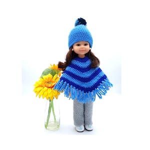 May include: A doll wearing a blue and white striped crocheted poncho with fringe, a blue crocheted hat with a pom pom, and grey crocheted trousers. The doll has brown hair and blue eyes.