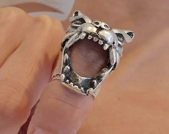 Op de afbeelding: Een zilverkleurige metalen ring met een sculptuur van een dierenkop met een open mond. De ring wordt om een vinger gedragen en toont gedetailleerde gelaatstrekken, waaronder puntige oren, een snuit en zichtbare tanden langs de open kaak.