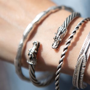 May include: A set of five silver bracelets, including two with dragon head accents, one with a feather design, and two plain bands.