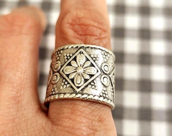 May include: A wide, silver-colored ring with an ornate floral and geometric design. The ring features a central diamond shape with a flower, surrounded by smaller circles and dots. The ring is on a finger with purple nail polish.