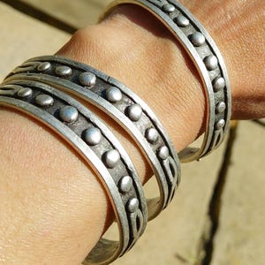 May include: Several silver-toned metal bracelets with a dotted design are stacked on a wrist. The bracelets have a curved shape and are worn on the arm. The bracelets are made of a shiny metal and are displayed in natural sunlight.