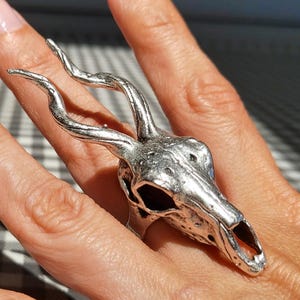 Könnte beinhalten: Ein silberfarbener Ring in Form eines Schädels mit geschwungenen Hörnern. Der Ring wird an einem Finger getragen und zeigt die komplizierten Details des Schädeldesigns. Ein einzigartiges Schmuckstück.
