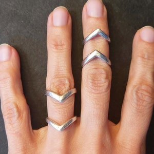 Könnte beinhalten: Drei silberne Chevron-Ringe, die auf einem Finger übereinander liegen. Die Ringe sind aus glattem Metall und haben ein schlichtes, modernes Design.