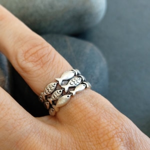 May include: A silver ring with a design of multiple fish stacked on top of each other. The ring is worn on a person's finger.