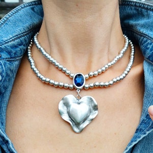 May include: A silver necklace with two strands of small beads. The necklace has a large, textured heart pendant and a blue oval gem. The necklace is worn over a denim jacket.
