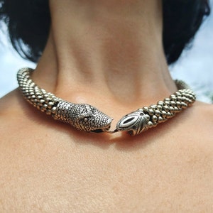 May include: A silver snake-shaped choker necklace with a textured finish. The necklace is made of metal beads and has a snake head clasp.
