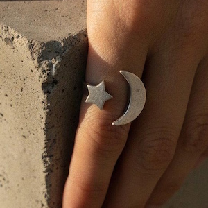 May include: A silver-toned open ring featuring a crescent moon motif on one end and a five-pointed star motif on the other end. The ring is designed with a gap between the two celestial shapes, allowing for an adjustable fit around the finger.