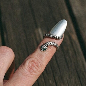 Puede incluir: Un anillo de serpiente plateado envuelve un dedo, con la cabeza de la serpiente cerca de la punta del dedo. El anillo tiene una textura similar a las escamas. El fondo es una superficie de madera.
