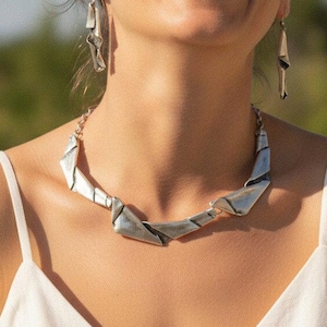 May include: A silver-toned metal necklace featuring angular, geometric links that lay flat against the collarbone, paired with matching dangling earrings. The jewelry set has a polished, reflective finish and a structured, modern design.