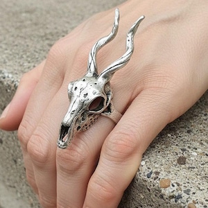 Puede incluir: Un anillo de tono plateado con un diseño detallado de cráneo de animal con cuernos largos y curvos. El metal tiene un acabado texturizado y desgastado con áreas oscuras hundidas que resaltan los rasgos anatómicos del cráneo y los cuernos.