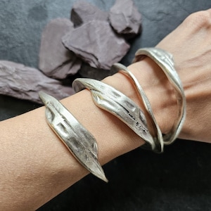 Pode incluir: Pulseira de punho em metal prateado com design de folha. A pulseira envolve o pulso, apresentando três faixas e um detalhe em forma de folha. A joia é exibida no braço de uma pessoa, com um fundo escuro.