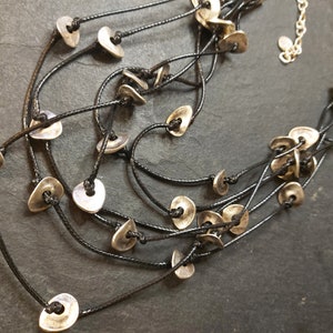 May include: A multi-strand necklace with silver-toned metal beads in the shape of a teardrop. The necklace is made of black cord and has a simple, minimalist design.