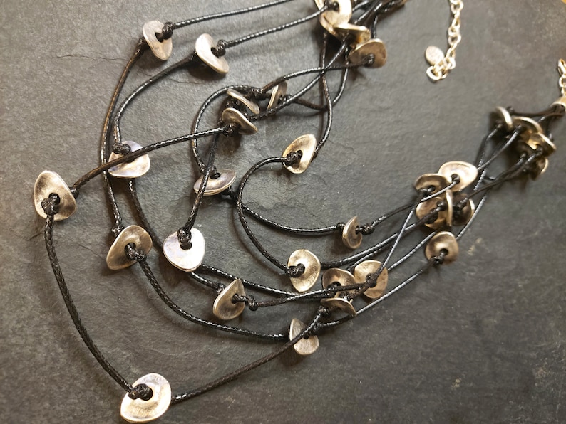 May include: A multi-strand necklace with silver-toned metal beads in the shape of a teardrop. The necklace is made of black cord and has a simple, minimalist design.