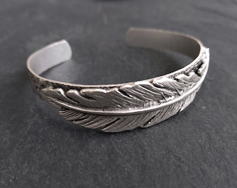 May include: A silver bracelet with a detailed feather design. The bracelet is made of metal and has a smooth, polished finish.