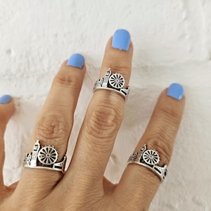 Puede incluir: Anillos plateados con diseño de noria, usados en dedos con esmalte de uñas azul claro. Los anillos tienen un diseño arquitectónico detallado, mostrando una noria y siluetas de edificios.
