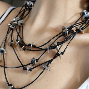 May include: A layered necklace with multiple strands of black cord and silver metal discs. The necklace is worn by a person, but the focus is on the necklace itself.