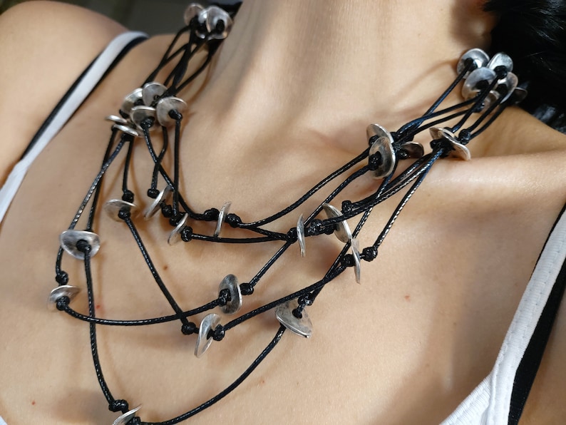 May include: A layered necklace with multiple strands of black cord and silver metal discs. The necklace is worn by a person, but the focus is on the necklace itself.