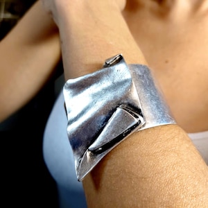 Puede incluir: Brazalete de puño plateado con un diseño texturizado y plegado. La banda ancha se envuelve alrededor de la muñeca, con una pieza de acento geométrica grande. El brazalete tiene un acabado de metal cepillado.