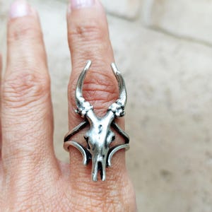 May include: A silver ring with a deer skull design. The ring has detailed antlers and facial features, with a band that encircles the finger. This distinctive ring is a unique piece of jewellery.