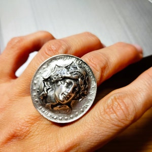 May include: A silver ring with a detailed design of a woman's face with a crown of leaves. The ring is worn on a finger.