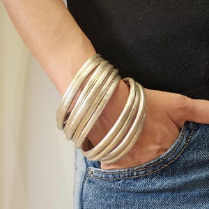 Op de afbeelding: Een stapel zilverkleurige armbanden om een pols. De armbanden zijn dik en hebben een getextureerd oppervlak. De persoon draagt een zwarte top en een blauwe spijkerbroek.