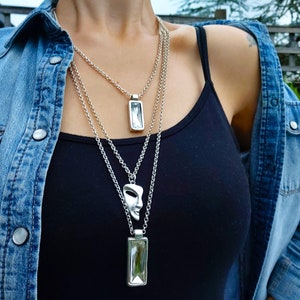 May include: A woman wearing a denim jacket and a black tank top with three silver necklaces. The necklaces feature a face pendant, a rectangular crystal pendant, and a smaller rectangular crystal pendant.