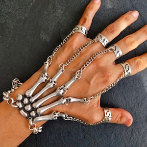 Puede incluir: Joyería de mano de esqueleto plateada. La pulsera presenta un diseño de mano esquelética con anillos para los dedos conectados por cadenas. Los anillos tienen un patrón decorativo recortado. La joyería se usa en una mano sobre un fondo oscuro.