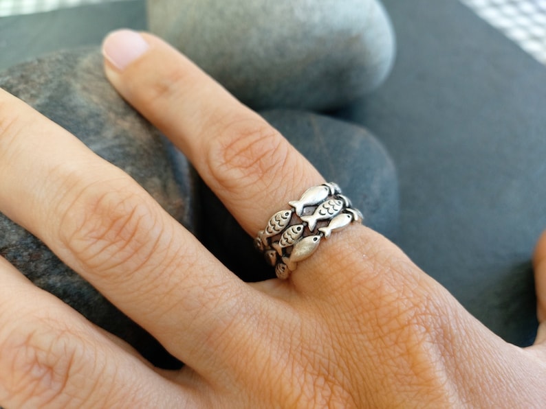May include: A silver ring with a design of multiple fish stacked on top of each other. The ring is worn on a person's finger.